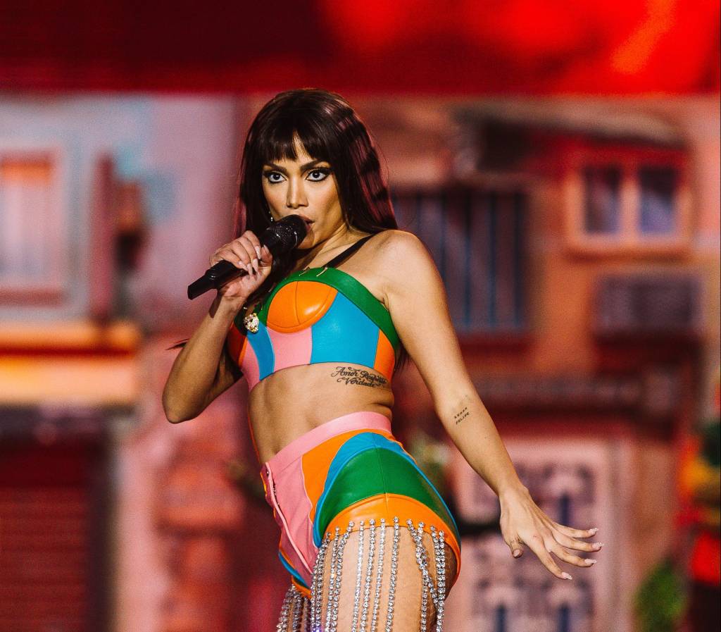 Anitta com top e hot pants nas cores verde, laranja, rosa e azul-turquesa. Ela está no palco cantando.