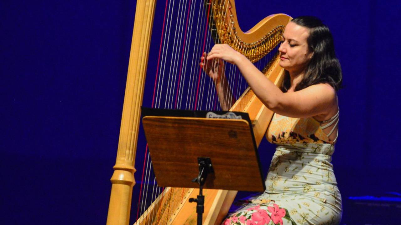 A harpista búlgara Diana Todorova é branca, tem cabelos pretos e lisos, usa vestido claro e está sentada tocando sua harpa. Ao fundo, uma luz azul-royal.