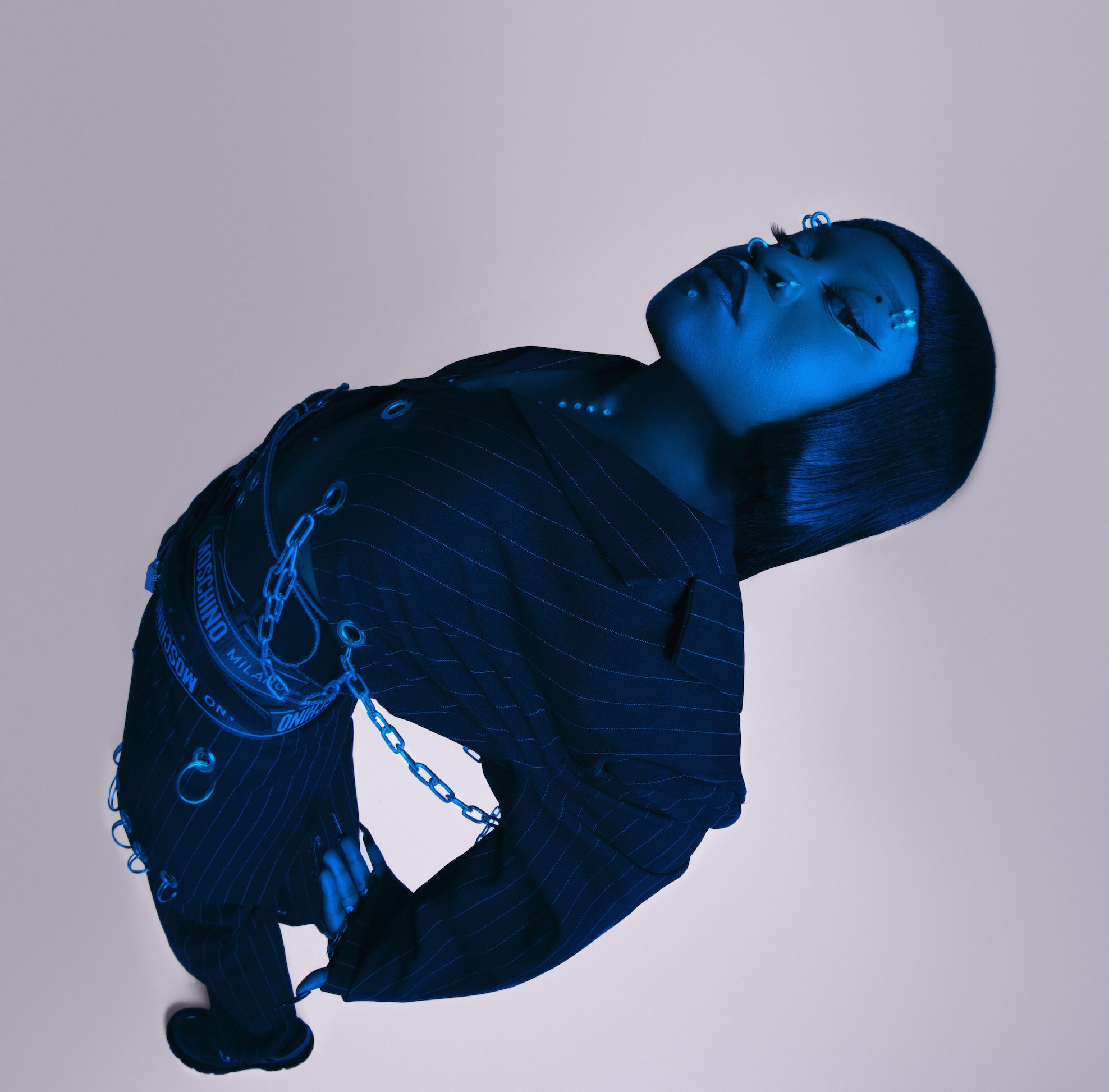 A cantora Sampa The Great é negra, está com os cabelos lisos, usa terno risca-de-giz e correntes e faixas com nomes de marcas como Moschino na cintura. A foto é tirada de cima, e ela está com as costas e os braços para trás. Uma luz azul deixa a artista com cor azulada.
