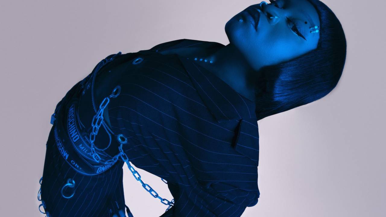 A cantora Sampa The Great é negra, está com os cabelos lisos, usa terno risca-de-giz e correntes e faixas com nomes de marcas como Moschino na cintura. A foto é tirada de cima, e ela está com as costas e os braços para trás. Uma luz azul deixa a artista com cor azulada.
