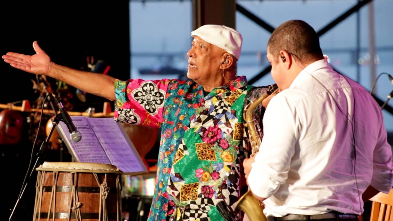 No centro da foto, o percussionista e compositor Carlos Negreiros. Ele é um homem negro, usa camisa estampada e boina branca. À direita, um saxofonista, branco, de costas.