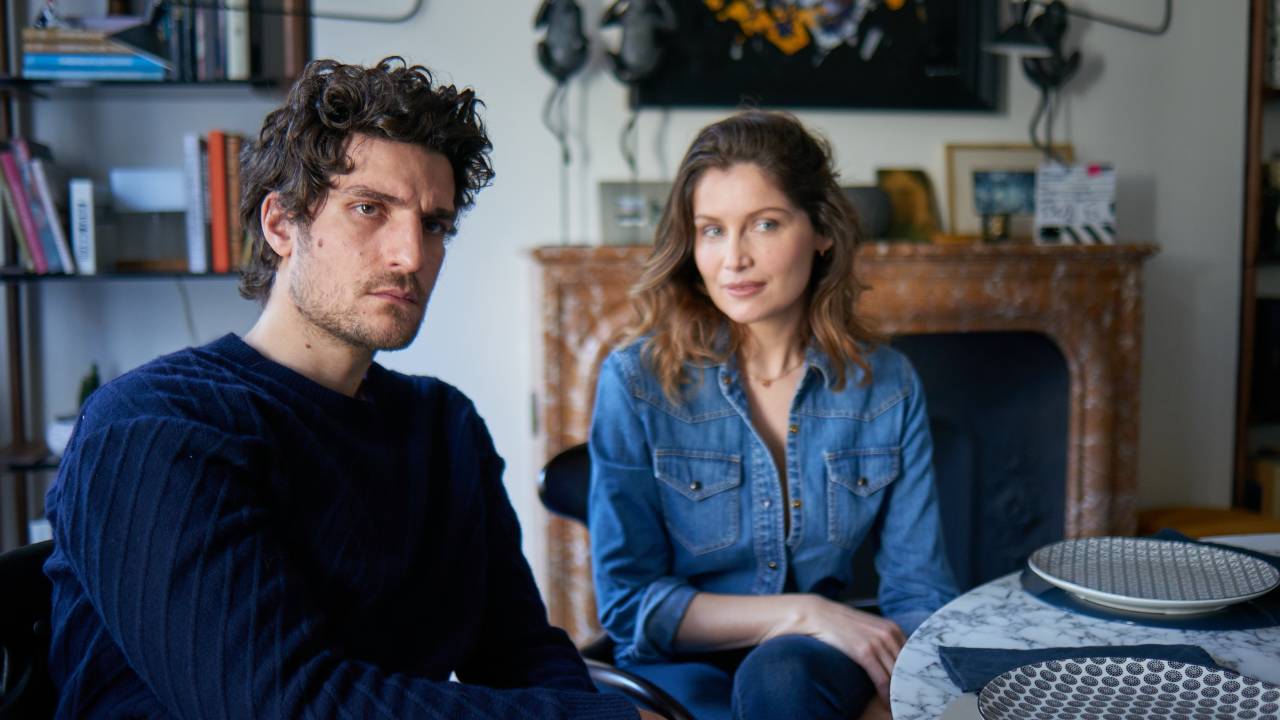 À esquerda, o ator Louis Garrel, de camisa azul-escuro de mangas compridas. À direita, a atriz Laetitia Casta, de vestido jeans. Eles estão sentados à mesa.