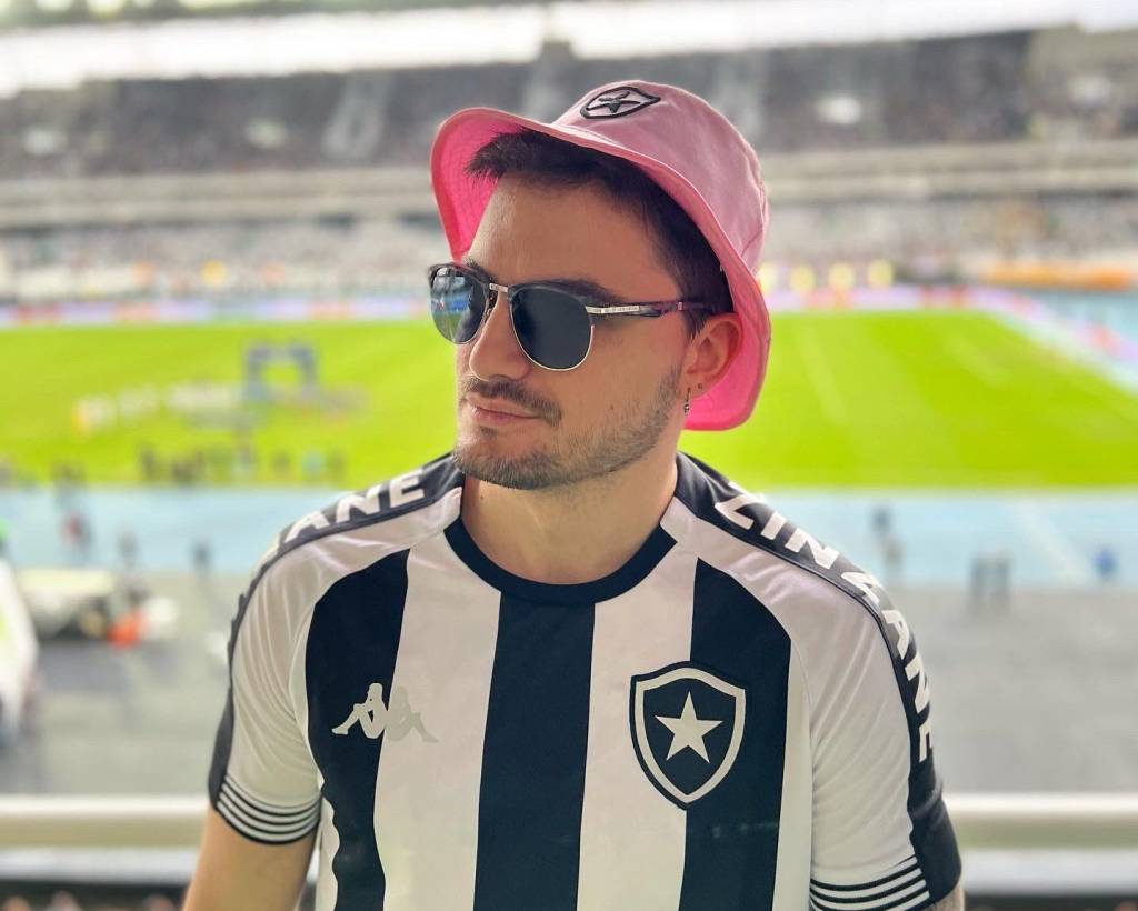 Felipe Neto usa camisa do Botafogo, chapéu rosa e óculos escuros. Ao fundo, o gramado do Engenhão.