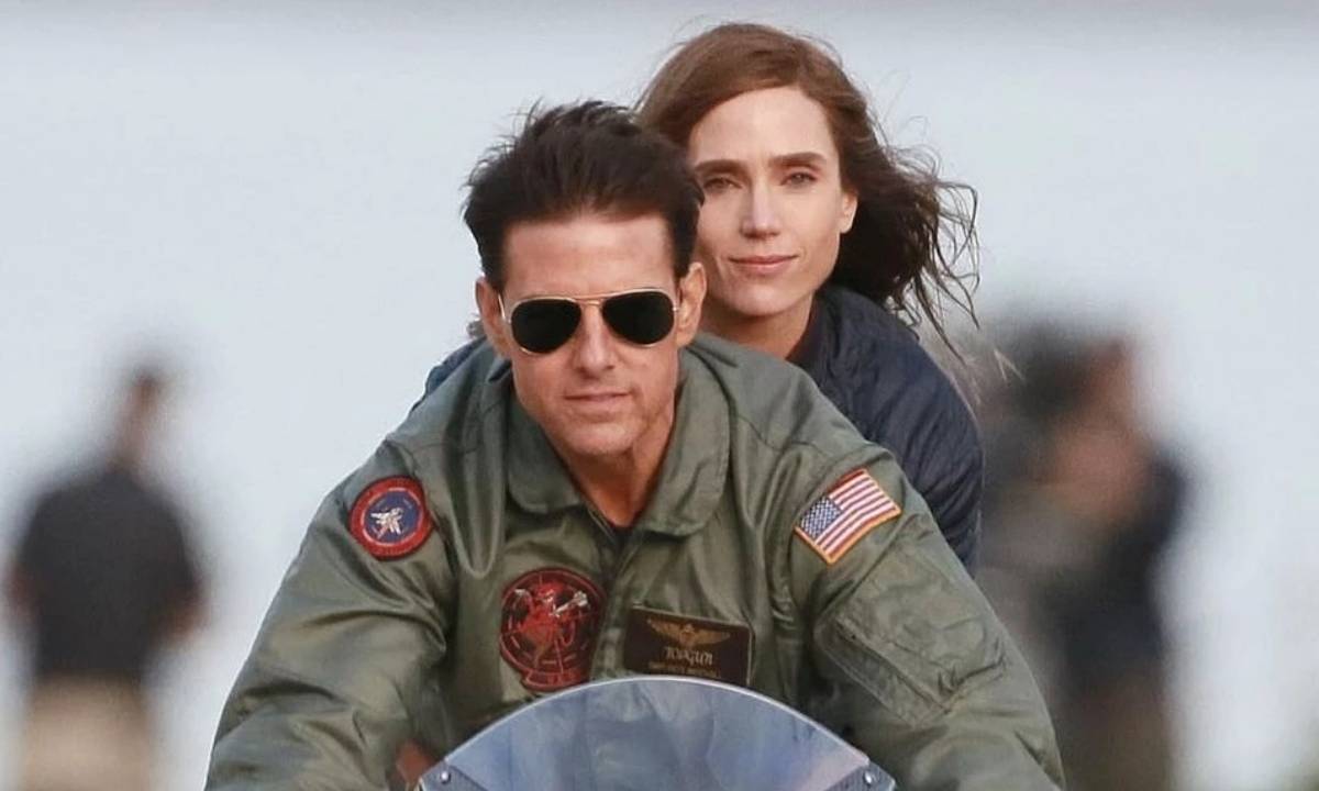 Tom Cruise pilota uma moto. Ele usa óculos escuros. Jennifer Connelly está sentada atrás dele.