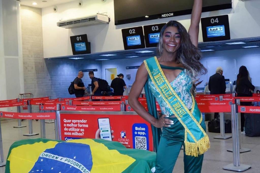 Foto mostra modelo trans em aeroporto usando uma faixa verde e amarela escrita "Miss T Brasil 2020"