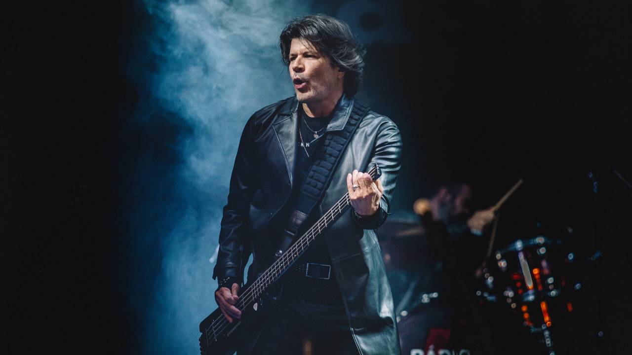 Paulo Ricardo está no palco, olhando para a esquerda da foto e tocando guitarra. Ele está vestindo roupa preta. Ao fundo, o baterista.