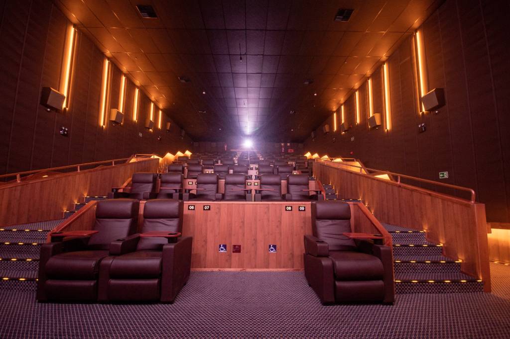Sala de cinema vista da primeira fileira, com a luz do projetor ao fundo, cadeiras marrons e detalhes de madeira.