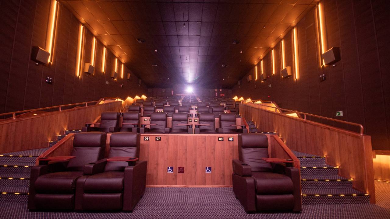 Sala de cinema vista da primeira fileira, com a luz do projetor ao fundo, cadeiras marrons e detalhes de madeira.