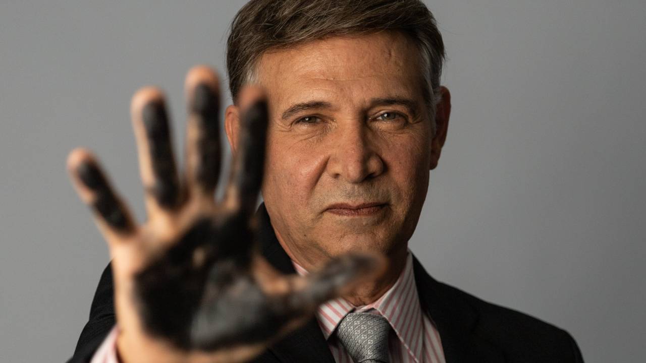 Nelson Freitas está representando Eike Batista. Ele usa peruca castanha com as laterais do cabelo grisalhas, além de terno preto com camisa branca e gravata cinza-claro. Está com a mão direita levanta e aberta em primeiro plano, com a palma das mãos e dos dedos suja de tinta preta.