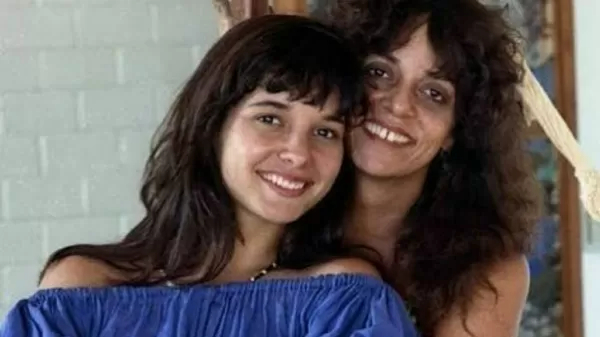 Daniella Perez está vestido blusa azul com decote ombro a ombro e é abraçada por trás pela mãe, Glória Perez. As duas estão olhando para a câmera e sorrindo.