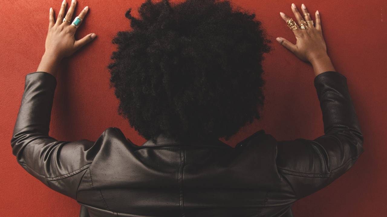Mulher negra com cabelo black power e jaqueta preta, de costas, com as mãos apoiadas na parede.