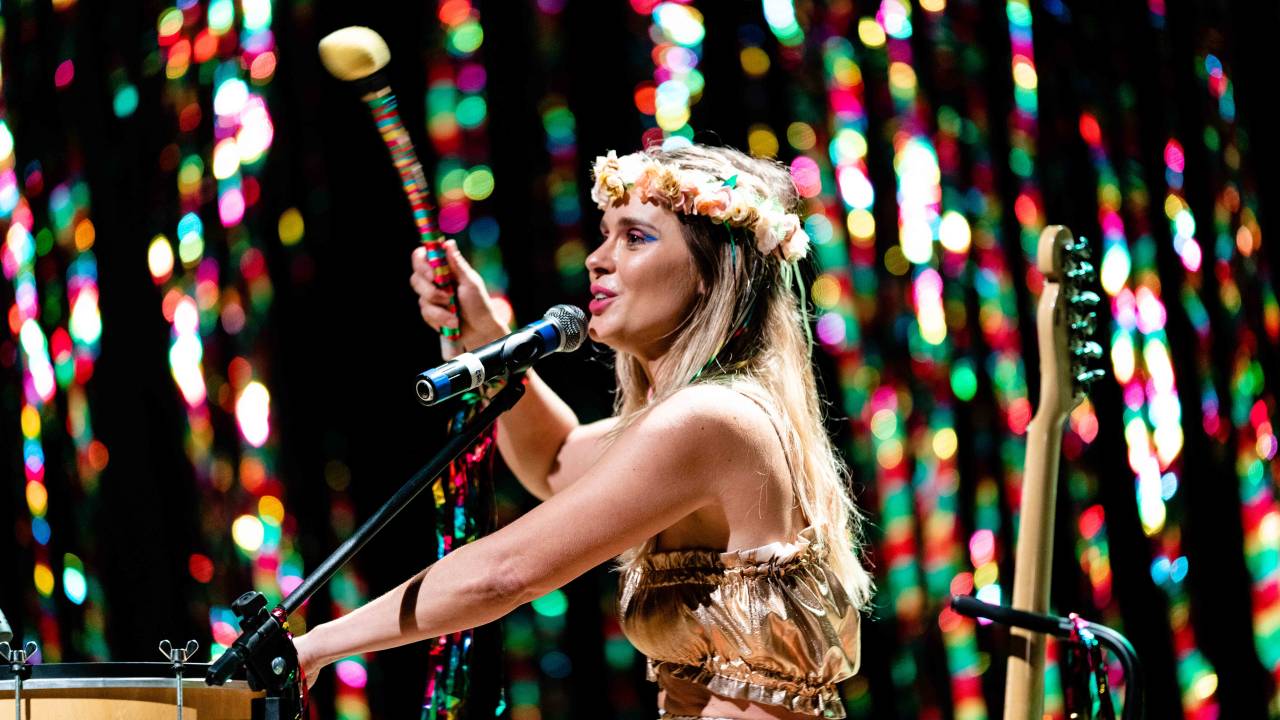 A foto mostra Carolina Dieckman de lado. Ela está de sentada, com o braço direito segurando uma baqueta de surdo no alto, cantando e tcando. Ela usa top e calça douradas e uma coroa de flores na cabeça.