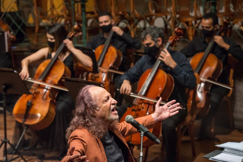 Alceu Valença de blusa preta e casaco marrom, tendo ao fundo os violoncelistas da Orquestra Ouro Preto.