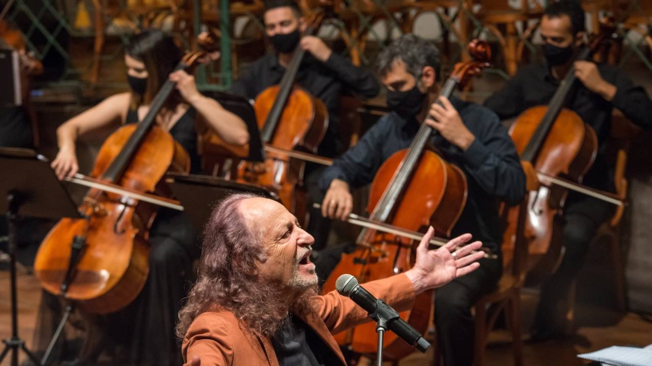 Alceu Valença de blusa preta e casaco marrom, tendo ao fundo os violoncelistas da Orquestra Ouro Preto.