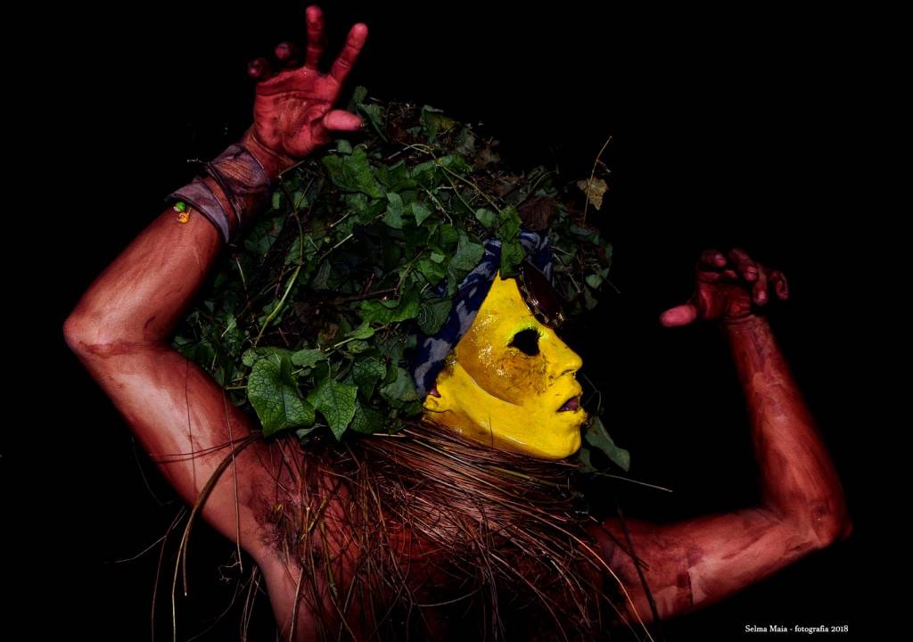 Fotografia Selma Maia com uma pessoa deitada de barriga para cima com folhas no lugar dos cabelos, tinta amarela no rosto e cipós em volta do pescoço.
