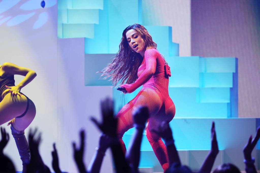 Foto de Anitta de lado, usando body vermelho, com o bumbum virado para o público. Em primeiro plano, desfocadas, as mãos do público para o alto.