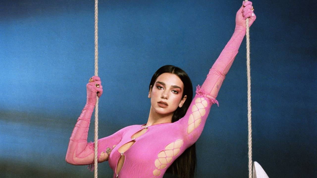 A cantora Dua Lipa de body rosa-choque e luvas na mesma cor, segurando as cordas de um balanço