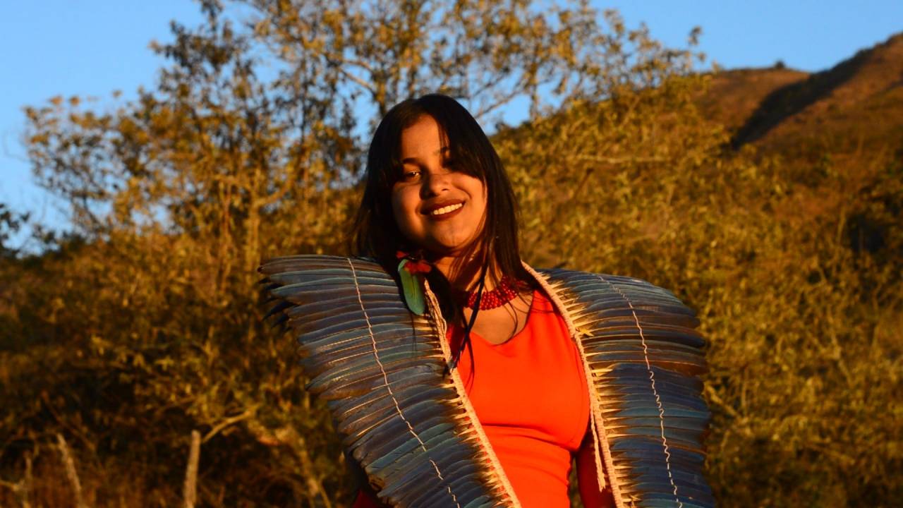 A cantora Kaê Guajajara de camisa coral e adereço de penas azul sobre os ombros