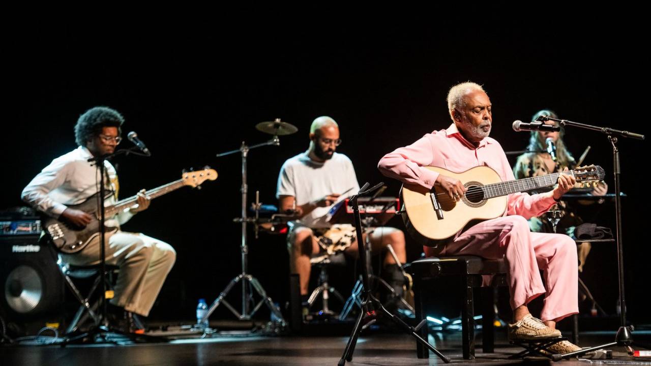 Gilberto Gil toca violão em show. Seus filhos Bem e José e a neta Flor aparecem ao fundo, tocando seus instrumentos.