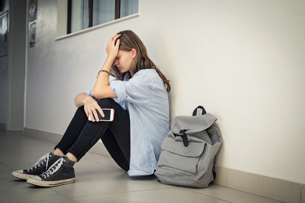 Adolescente sentada no chão leva a mão à cabeça, angustiada.