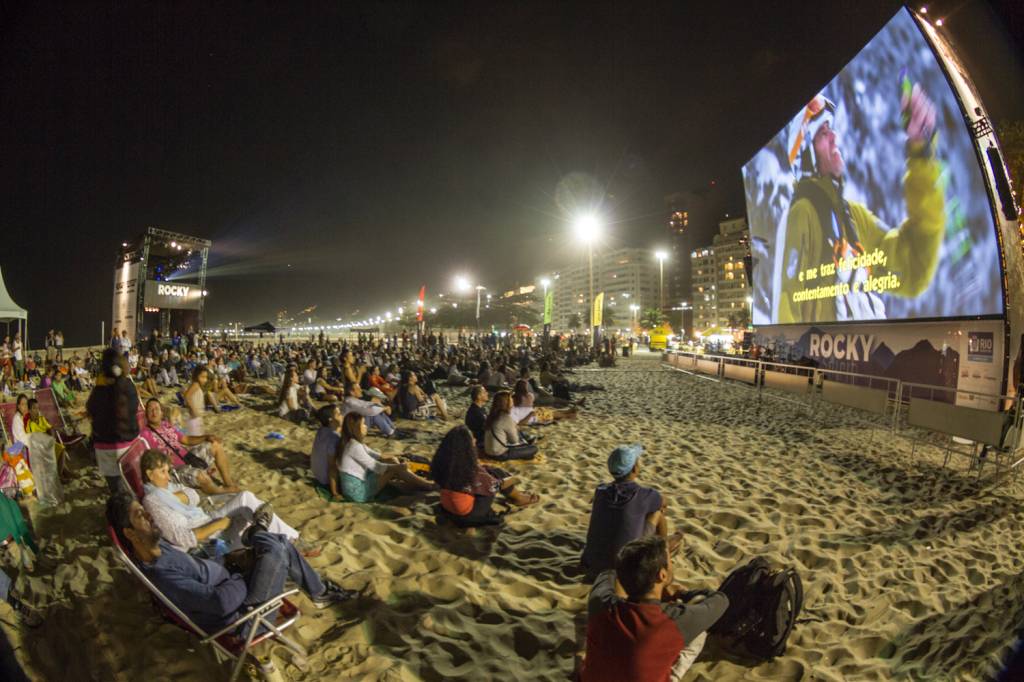 Foto mostra telão exibindo filme na praia de Ipanema à noite, com público sentado na areia assistindo