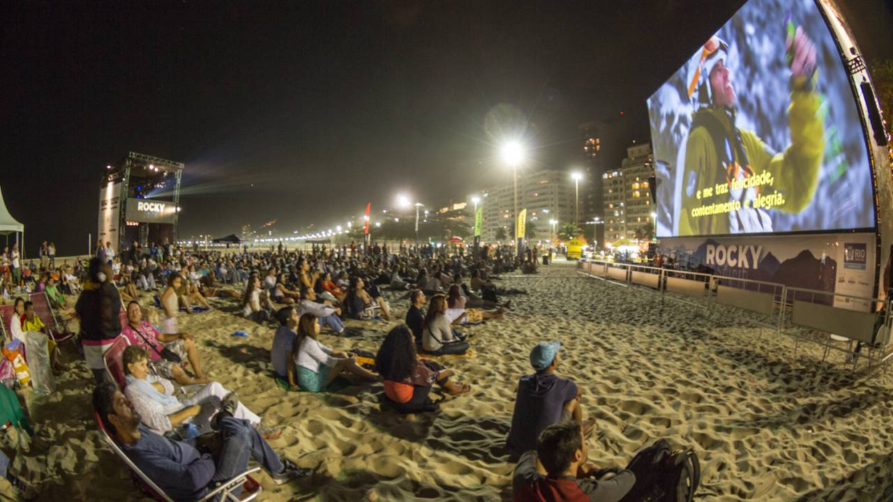 Foto mostra telão exibindo filme na praia de Ipanema à noite, com público sentado na areia assistindo