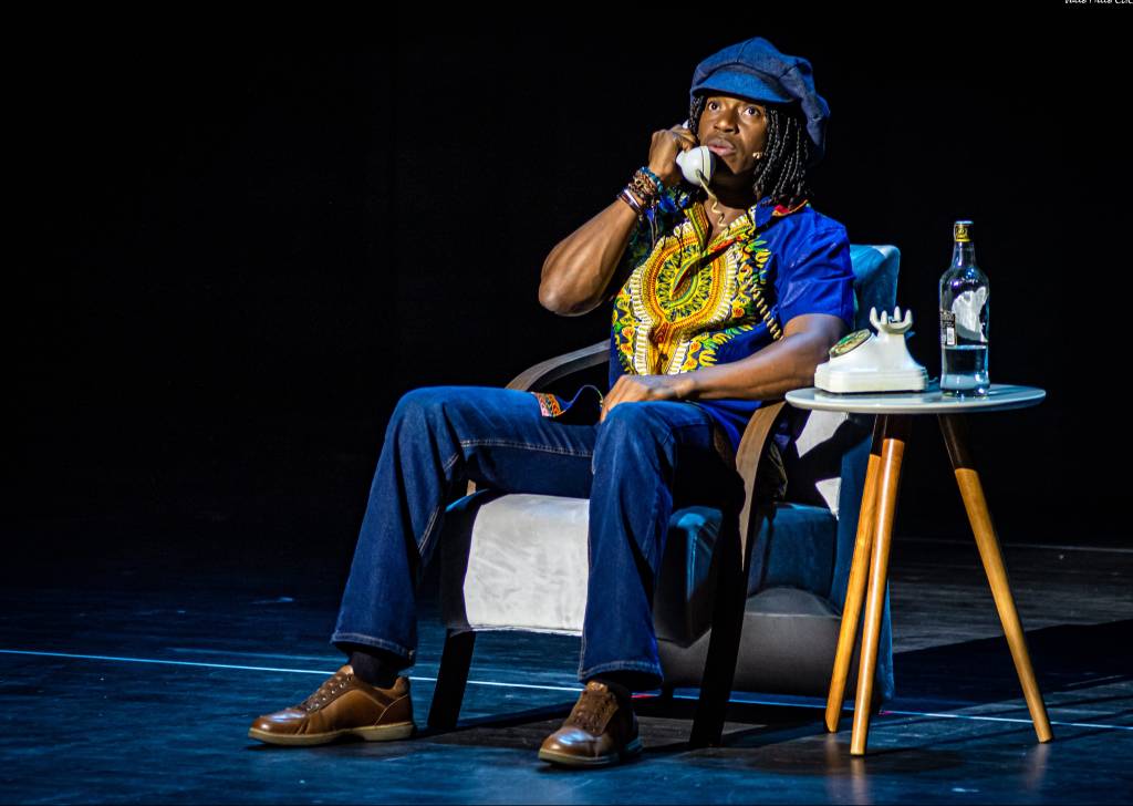 Tiago Barbosa caracterizado como Milton Nascimento, com boina azul-escuro, sentado em uma poltrona e falando ao telefone