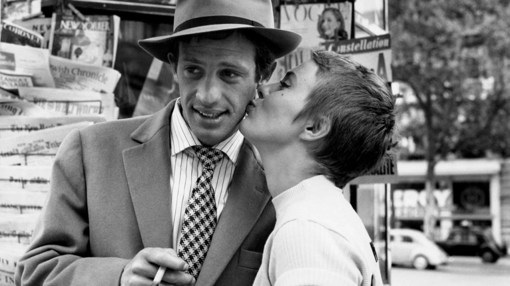 Foto mostra cena de filme de Godard em que mulher dá beijo na bochecha de um homem usando chapéu e terno, segurando um cigarro na mão