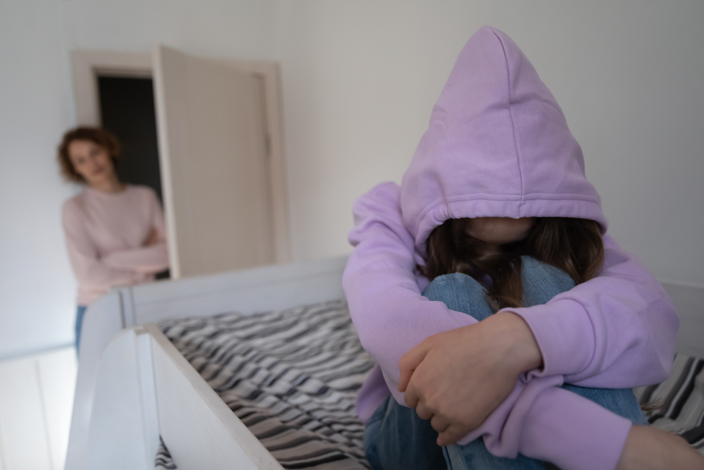 Adolescente esconde a cara sob um capuz e é observado pela mãe.