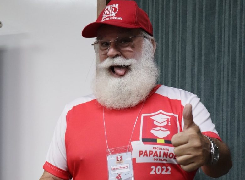 Homem de cabelo, barba e bigodes brancos, com blusa vermelha com o nome da Escola de Papai Noel do Brasil e e mangas brancas, usando boné vermelho e fazendo sinal de positivo.