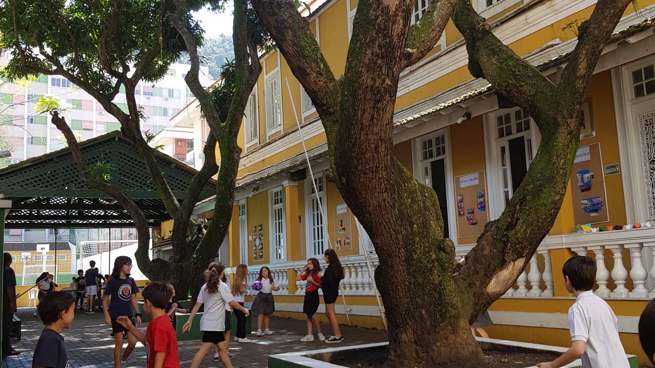 Fachada do prédio hisórico do colégio, amarela com detalhes brancos, com as crianças brincando no pátio arborizado.