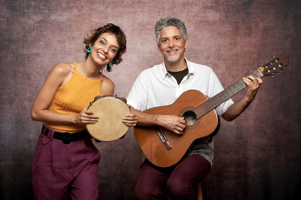 Foto mostra mulher segurando pandeiro e sorrindo ao lado de homem segurando violão e sorrindo