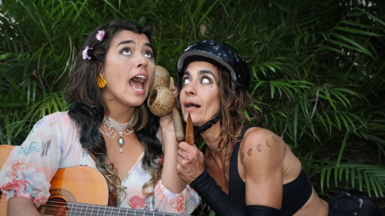 Foto mostra duas atrizes cantando. Uma delas um toca violão