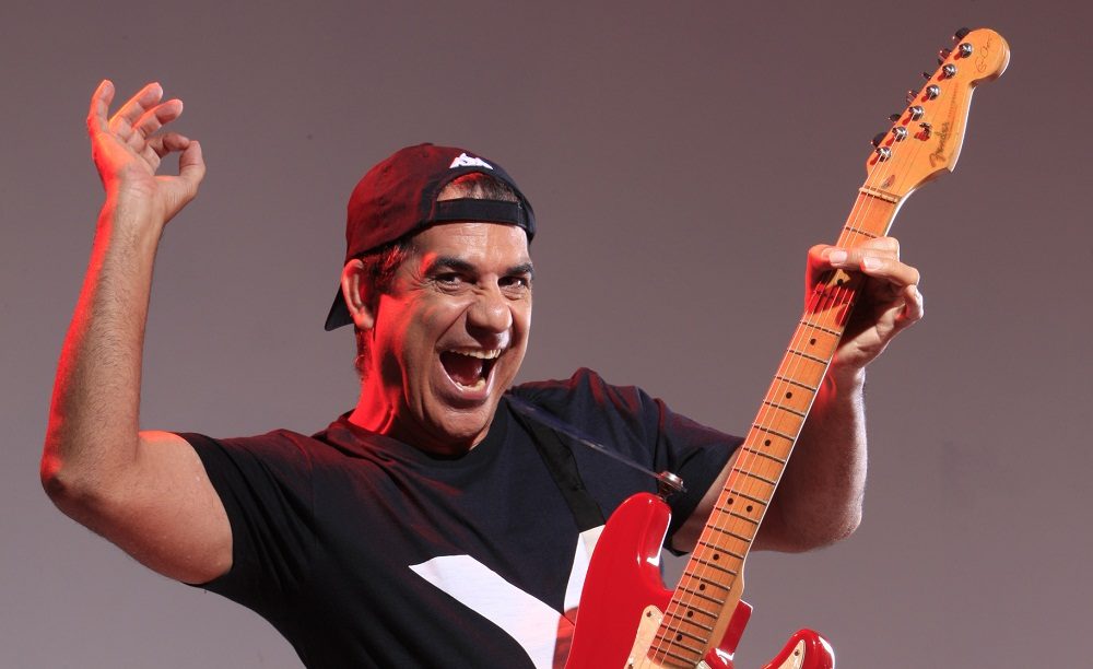 Durval Lelys com camiseta preta e boné virado para trás, segurando uma guitarra vermelha para o alto e com o braço direito também para o alto, como se estivesse tocando o instrumento.