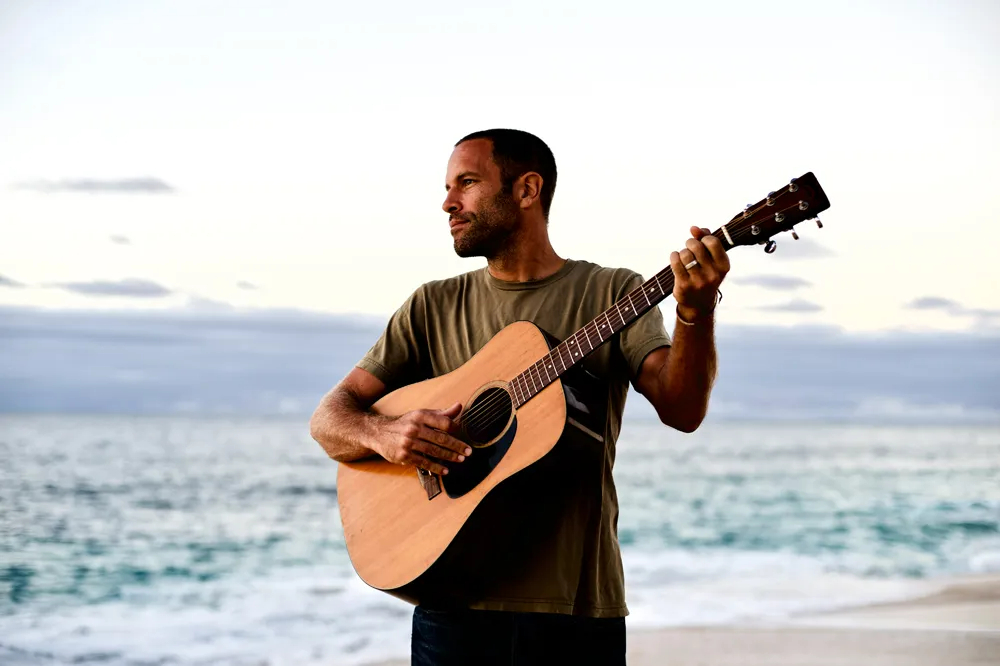 Jack Johnson de pé, tocando violão em uma praia, olhando para o lado esquerdo da foto