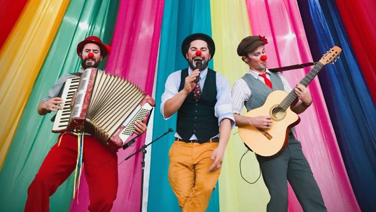 Foto mostra trio de homens com nariz de palhaço tocando música