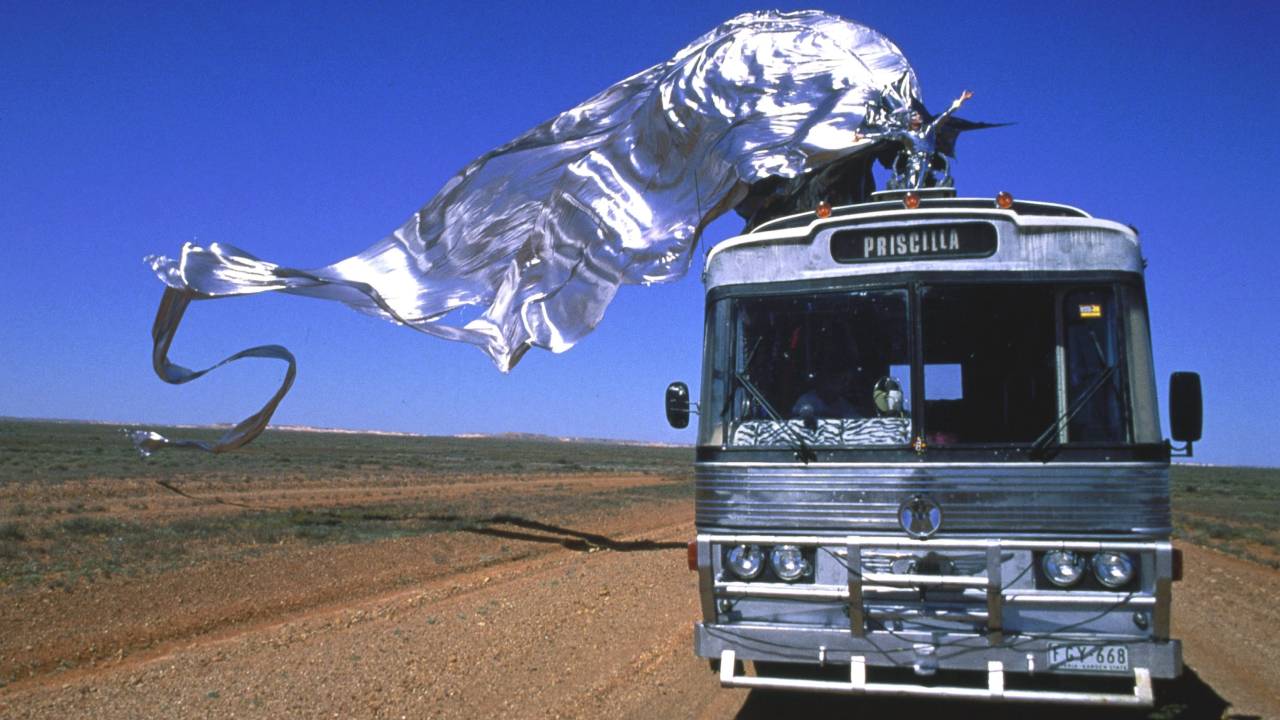 Cena do filme em que um ônibus prateado está no deserto e uma drag com roupa prateada está com uma longa capa esvoaçante.