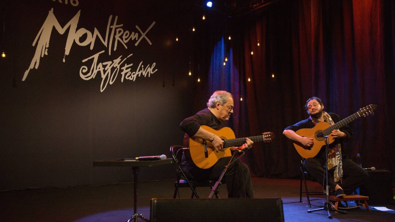 Toquinho e Yamandu Costa sentados tocando violão. Ao fundo, o letreiro do Rio Montreux Jazz Festival.