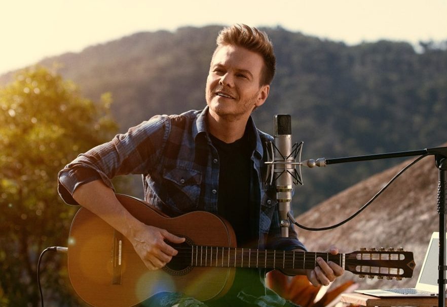 Michel Teló sentado, tocando violão, usando blusa preta e camisa jeans, em uma paisagem bucólica.