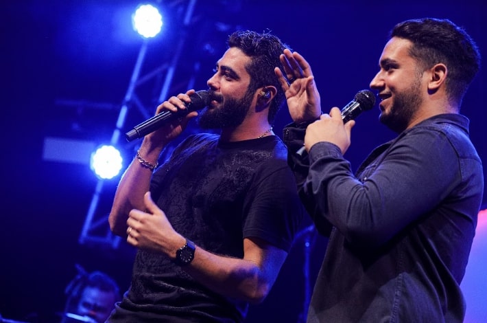 Os irmãos Henrique & Juliano de perfil, no palco, cantando, sob luzes azuis