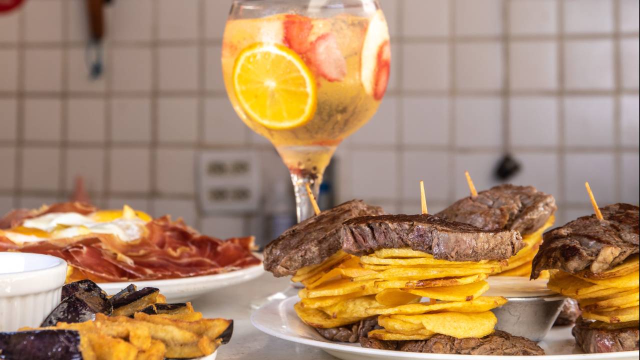 Foto mostra mesa com drinque, carnes e batata frita