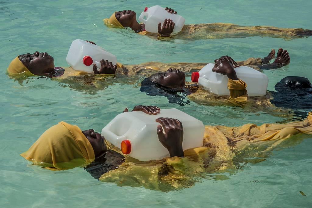 Foto mostra meninas aprendendo a nadar com toucas e roupas de banho amarelas. Elas boiam enquanto seguram um galão de água no peito