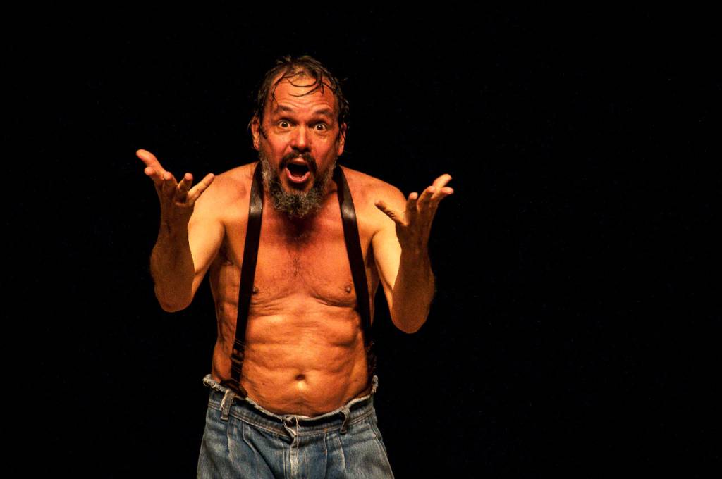 Foto mostra ator sem camisa usando uma calça e suspensórios. Ele está diante de um fundo preto, com os cabelos molhados e barba longa, e aponta as mãos em direção à tela