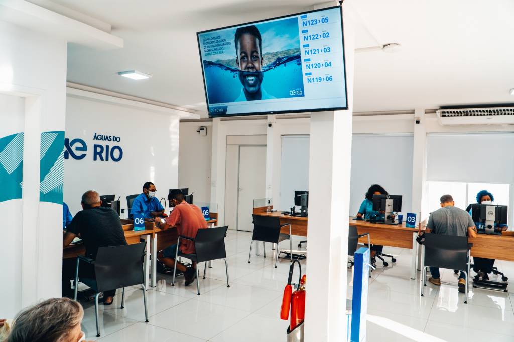 Foto mostra loja da Águas do Rio. Há quatro atendentes sentados em frente a computadores. Eles atendem os clientes. Na sala, toda na cor branca, há uma televisão que passa conteúdos referentes à concessionária