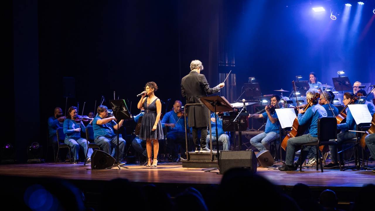 Foto mostra orquestra sinfônica e cantora se apresentando no palco