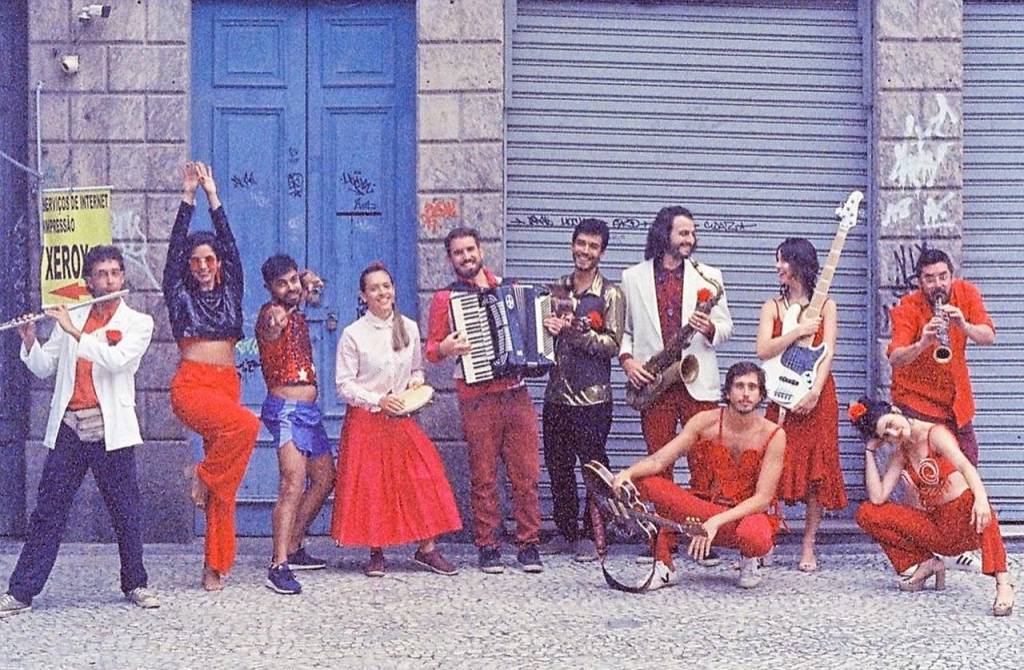 Os onze músicos, com peças de roupa vermelhas na frente de um sobrado antigo