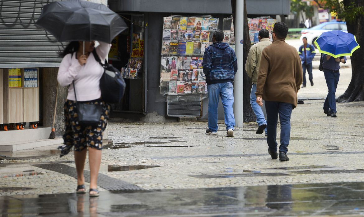 Foto mostra pessoas andando em rua do Rio com chuva. Uma mulher passa com um guarda-chuva e outros dois homens andam na direção oposta em frente a uma banca de jornal com revistas