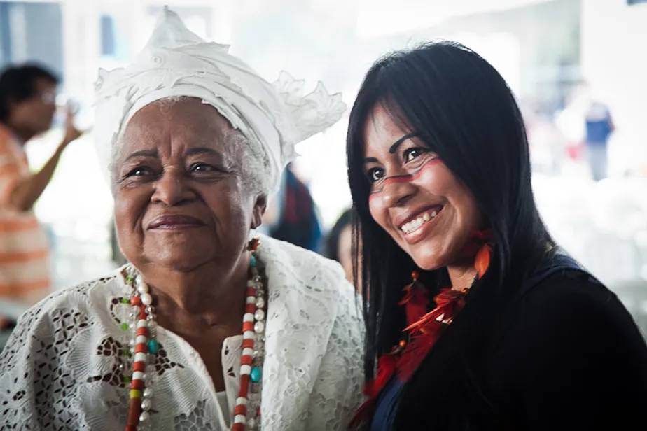 Foto mostra mulher jovem indígena e outra senhora com trajes religiosos de matriz africana