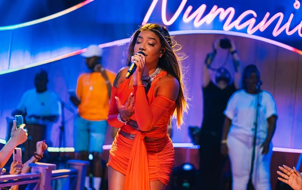 Foto mostra cantora Ludmilla usando vestido laranja e cantando com microfone