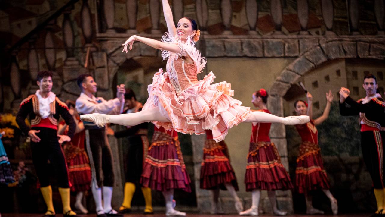 Em primeiro plano, a bailarina dando um salto com as pernas em espacate. Ao fundo, uma fileira com três bailarinos, quatro bailarinas e mais um bailarino com roupas que lembram as de dançarinos de flamenco.